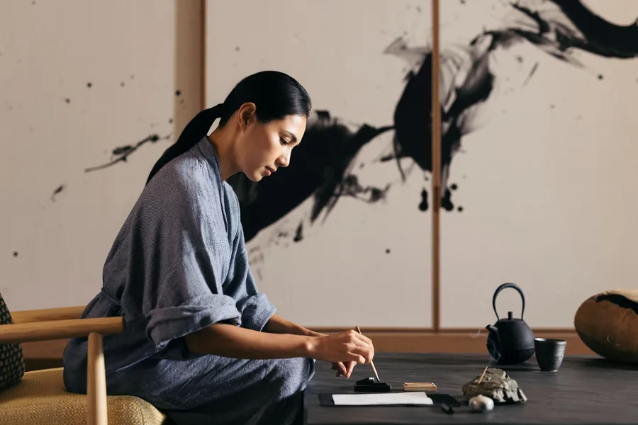 Ein Gast bereitet an einem niedrigen Tisch neben einer Teekanne Tusche für die Kalligrafie vor, eine japanisch geprägte Retreat-Szene von Six Senses